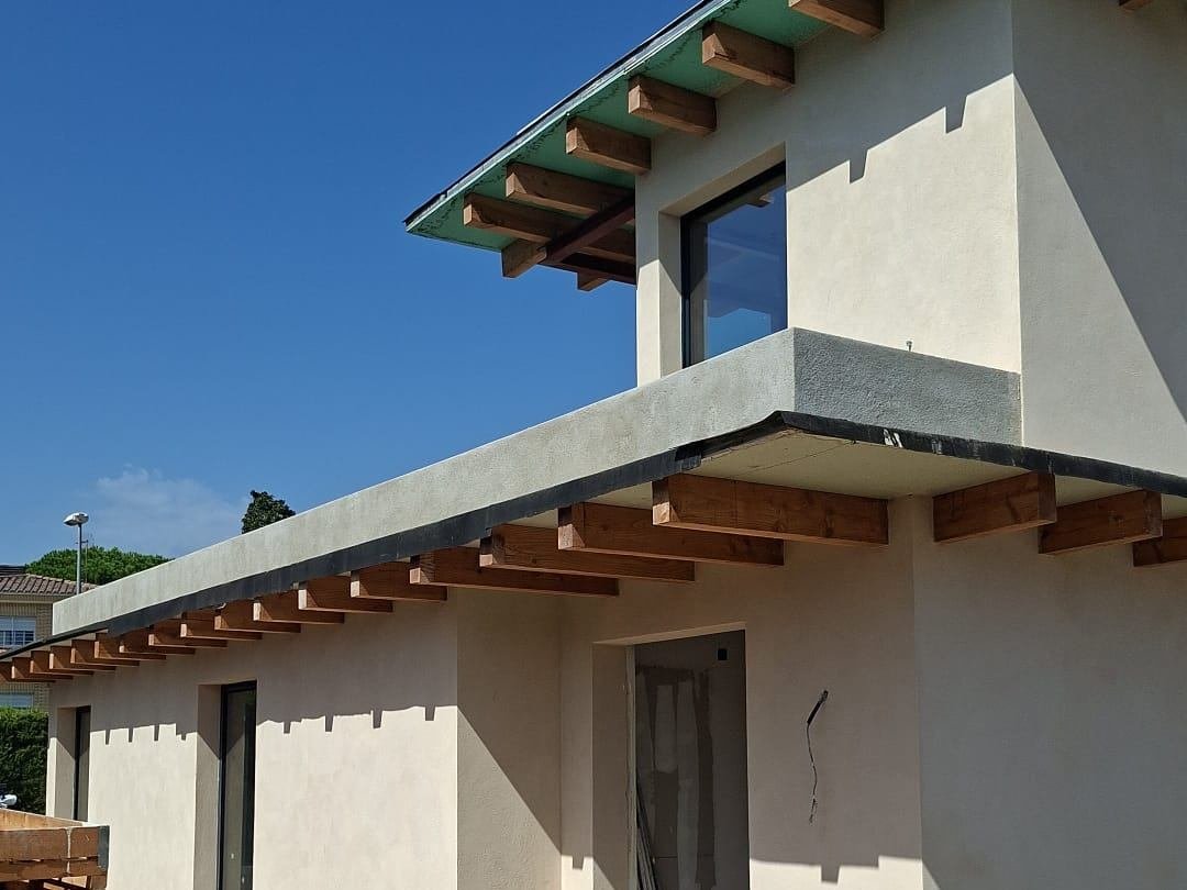 Vivienda unifamiliar en El Masnou con la fachada acabada en revoco de cal en tono arena, aleros con las vigas de madera vistas y bandas de hormigón bajo el forjado