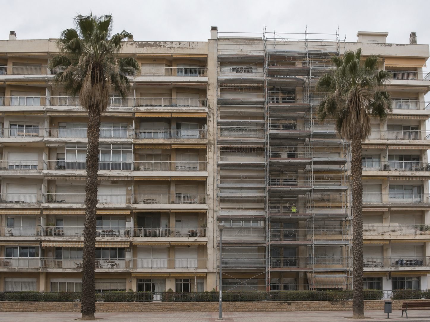 Gran bloque de apartamentos de la costa de Tarragona con largas hileras de balcones, andamio montado en una sección de la fachada y dos palmeras por delante