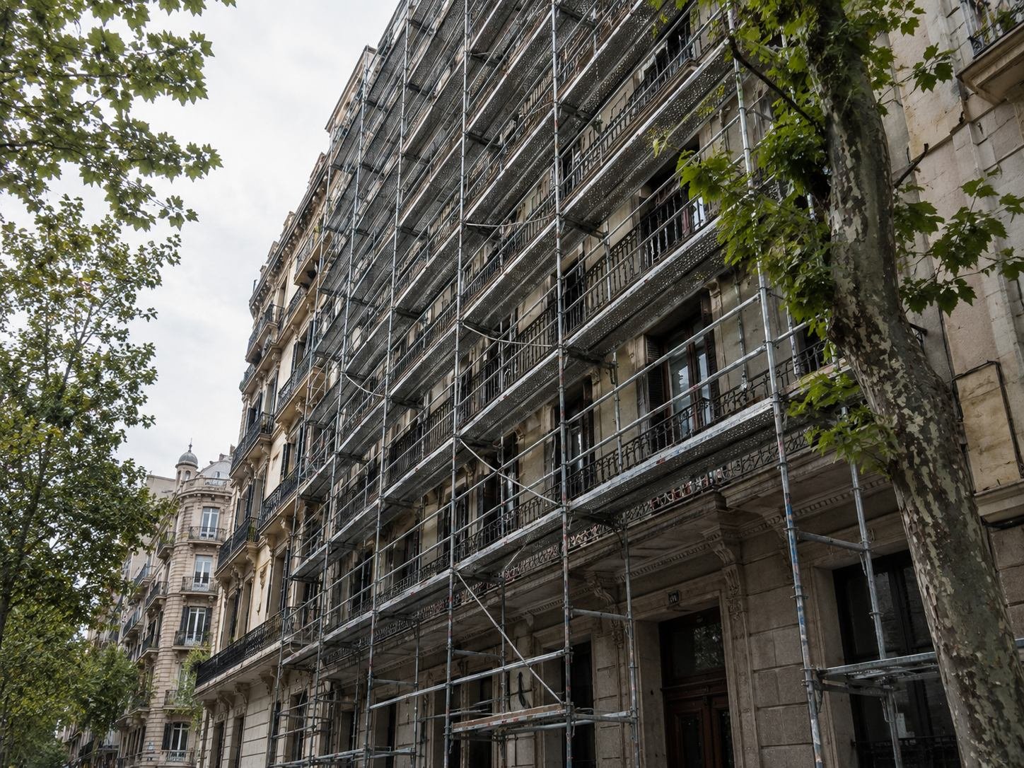 Andamio montado sobre la fachada de un edificio del Eixample de Barcelona, visto en escorzo desde la calle arbolada, con los balcones de forja detrás de la estructura