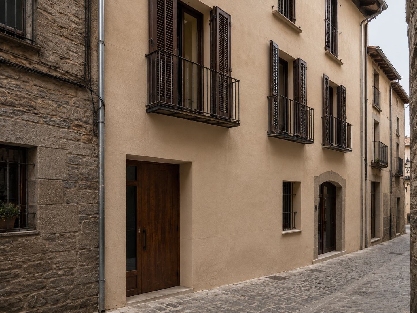 Fachada rehabilitada con estuco a la cal en tono tierra, en una calle empedrada y estrecha del casco antiguo, junto a un edificio de piedra vista