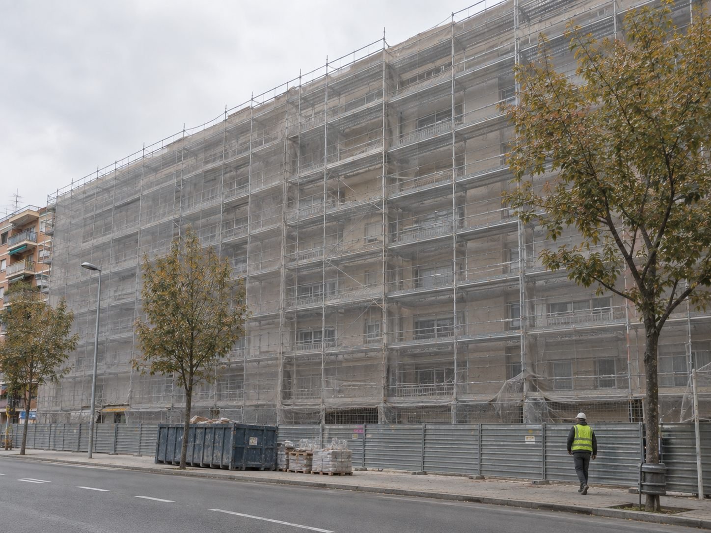 Bloque de viviendas grande y alargado en una avenida de Sabadell, cubierto de andamio, con un contenedor de obra y valla peatonal en la acera