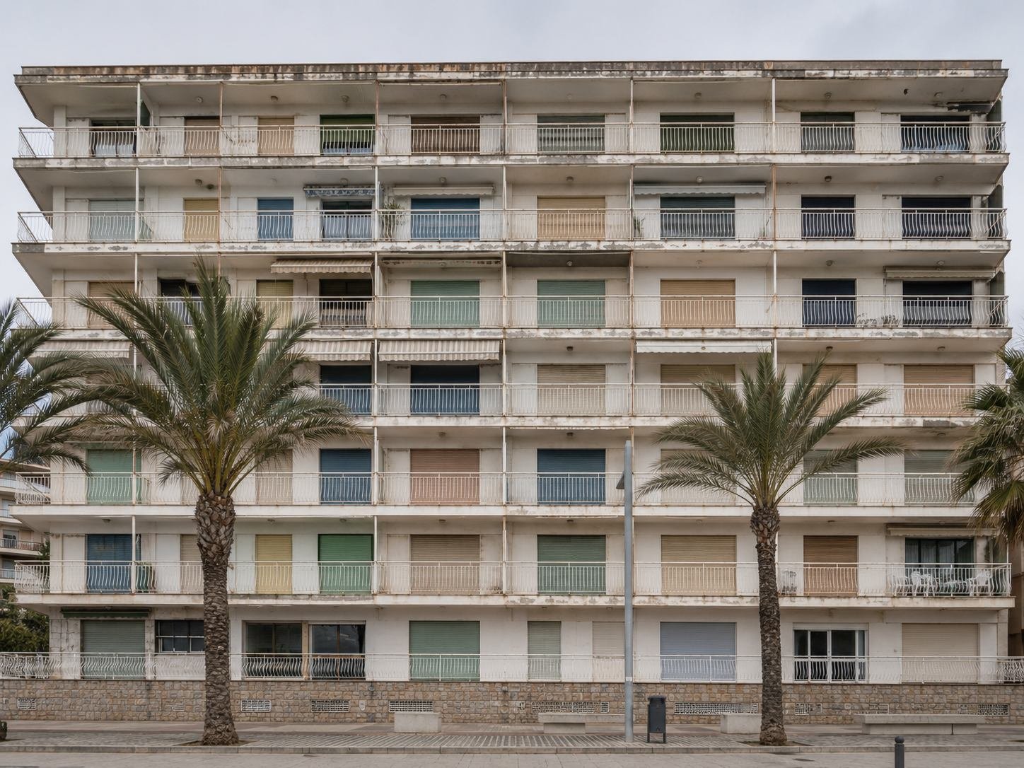 Bloque de apartamentos de los años setenta en primera línea de costa, con galerías de balcones corridos, persianas descoloridas y palmeras en primer término
